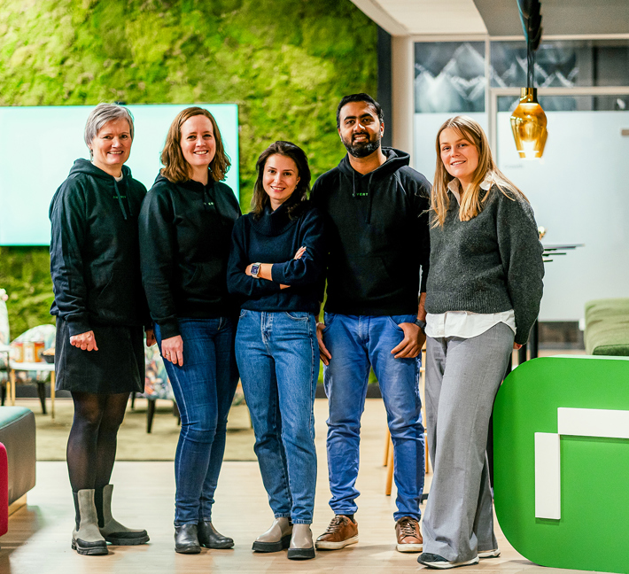 A diverse group of five employees standing together in a modern office environment, smiling towards the camera. From left to right, there's a woman in a black dress and boots, a woman in a black hoodie and jeans, another in a dark sweater and blue jeans, a man in a black hoodie and blue jeans, and a woman in a gray sweater and light gray pants. They are standing in front of a large green 'R' symbol and a screen with a green wall in the background, indicating a vibrant workspace.