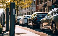 A row of electric vehicles parked at charging stations along a city street on a sunny day, with buildings and trees in the background.