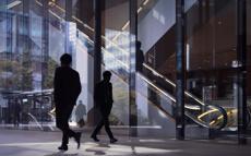 Two men in suits walk past each other in front of a modern glass building.