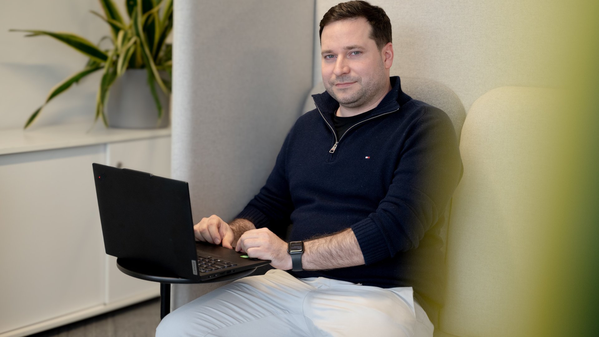A person sits in a high-backed, cushioned office booth, using a black laptop placed on a small round table. They are wearing a dark navy zip-collar sweater, light-colored pants, and a smartwatch. A potted plant with long green leaves sits on a cabinet in the background, suggesting a modern, quiet workspace designed for focused work