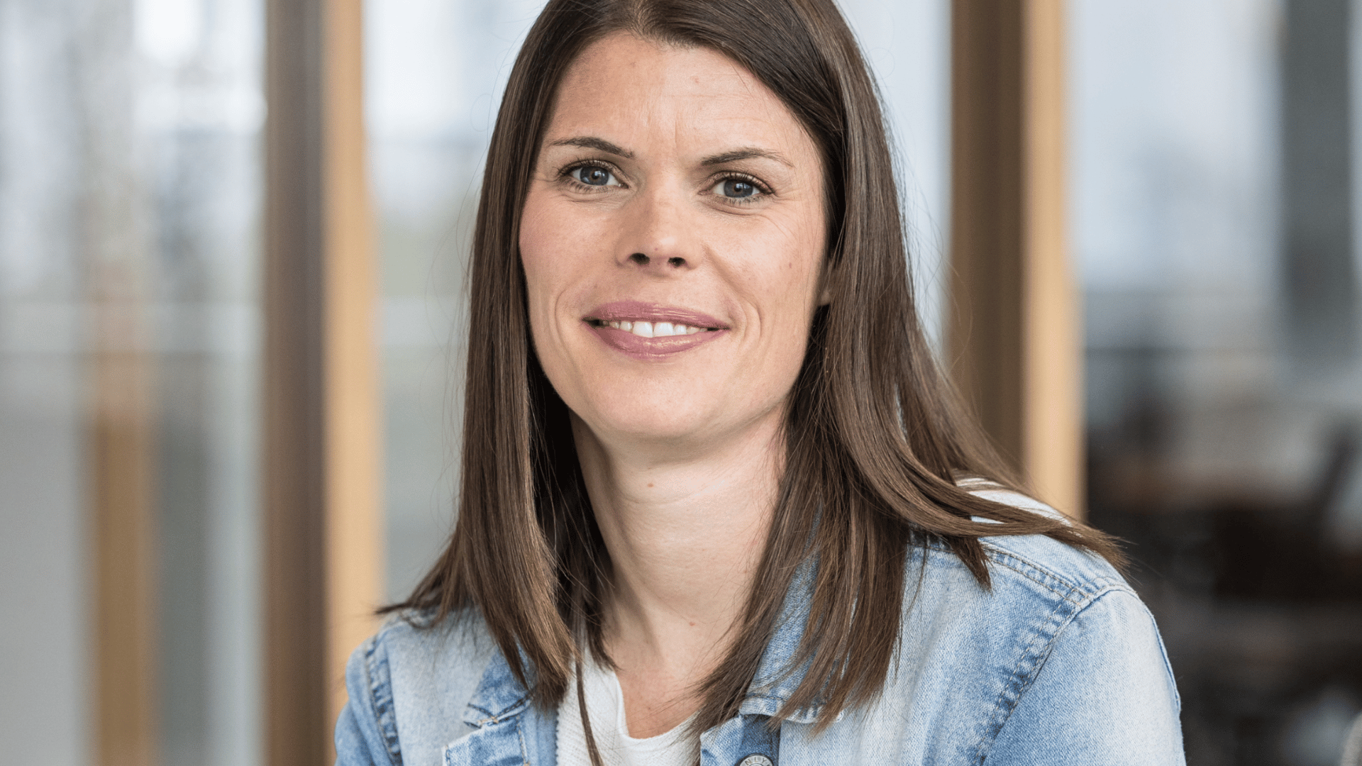 Circular portrait photo of a person with long brown hair and a friendly smile. Denim jacket, light background.