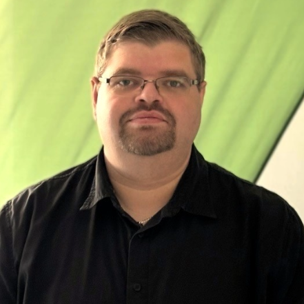 A person is standing in front of a semi-translucent green and white background that resembles a modern fabric banner or sail. They are wearing a black collared shirt and thin metal-framed glasses. The person has short brown hair, a neatly trimmed goatee, and is looking directly at the camera with a calm, neutral expression. Soft lighting comes from the left side, casting gentle shadows. No brand elements, technological devices, or accessibility-related symbols are visible.