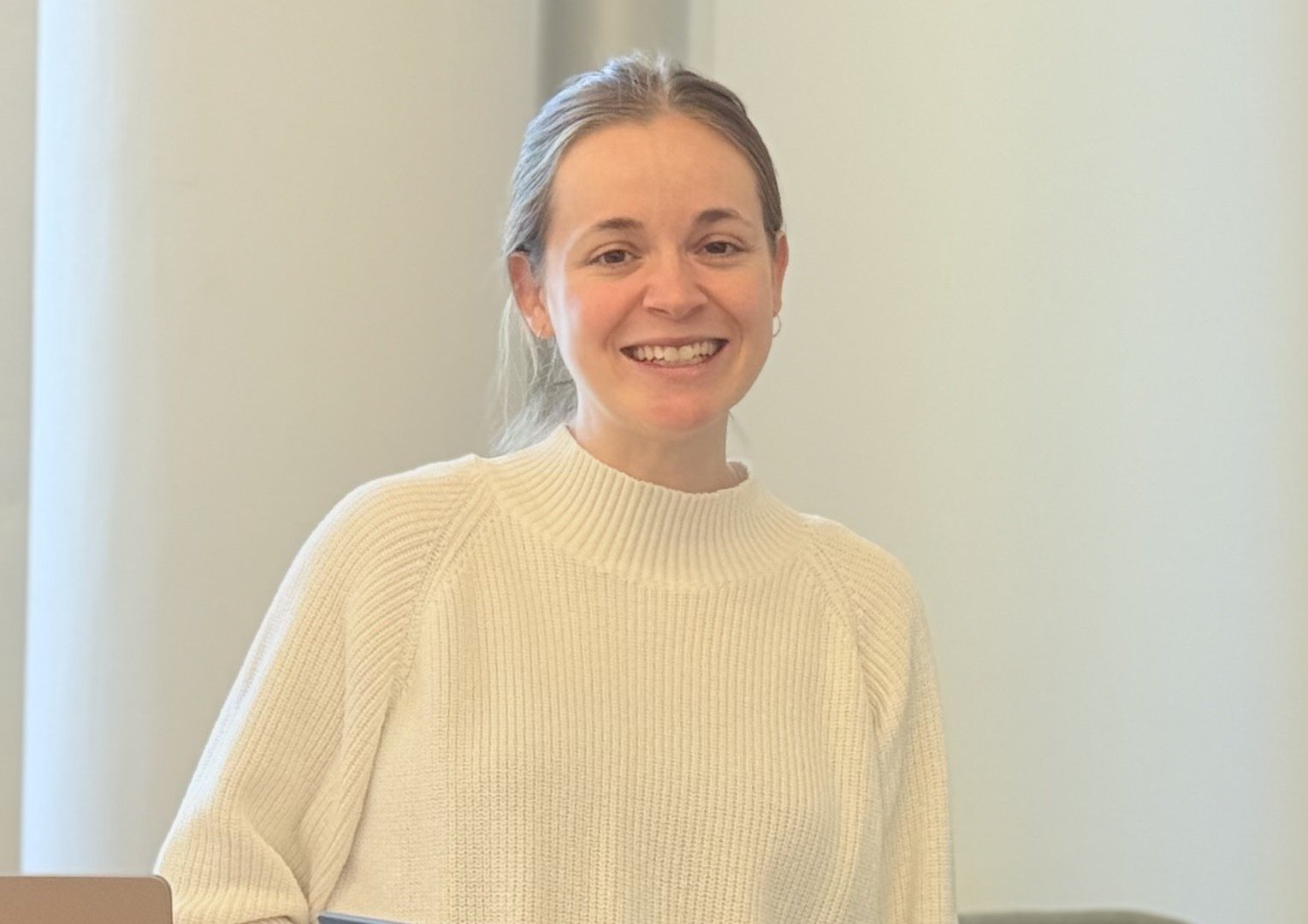 Eine lächelnde Person mit zurückgebundenem, hellem Haar steht in einem Innenraum und hält einen Laptop in der Hand. Sie trägt einen cremefarbenen Pullover, und weiches natürliches Licht fällt von der linken Seite ins Bild.