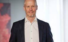 Porträtt av en man i mörk kavaj och vit skjorta, stående inomhus.