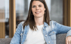 The image shows Carina seated in a relaxed posture on a grey upholstered sofa or armchair. The person has long, straight, dark brown hair worn open, falling onto their shoulders. Their facial expression is friendly and open: they are smiling naturally into the camera, conveying approachability and ease. The person is wearing a white top with a fine texture and a light blue denim jacket over it. Small green earrings are visible as accessories. One arm rests loosely on the back of the seat, emphasising the relaxed body posture. The background shows a modern, bright office or workspace with large windows through which natural daylight enters. The environment appears professional and welcoming. No text, logos, technologies, or accessibility symbols are visible in the image. The photograph is composed as a professional portrait, likely intended for a professional or institutional context.
