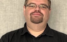 Eine Person steht vor einem neutralen, strukturierten grauen Hintergrund. Die Person trägt ein kurzärmliges, schwarzes Hemd mit Knopfleiste und eine dunkle Smartwatch am linken Handgelenk. Die Arme sind vor dem Körper locker verschränkt. Die Person hat kurzes, braunes Haar, trägt eine Brille mit schmalem Metallrahmen und einen gepflegten Kinnbart. Der Gesichtsausdruck wirkt ruhig und freundlich. Es sind keine marken- oder barrierebezogenen Elemente im Bild sichtbar.