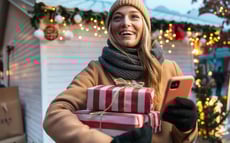 Junge Frau auf einem weihnachtlichen Markt, die lächelnd Geschenke trägt und ihr Smartphone benutzt