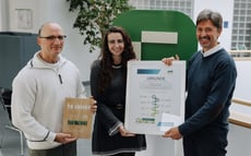 Drei Personen stehen in einem Innenraum und halten zwei Auszeichnungen. Die Person links hält eine Holzplakette zum zehnjährigen Jubiläum der Partnerschaft, während die beiden Personen in der Mitte und rechts eine gerahmte Urkunde präsentieren. Im Hintergrund ist ein großes grünes Riverty-Logo zu sehen.