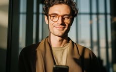 A young man using his smartphone in a bright modern office, smiling confidently in the sunlight.