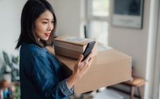 A woman carrying two cardboard packages in her left arm is looking at her smartphone while standing indoors. She is wearing a blue jacket and has a slight smile on her face. In the background, a softly lit room is visible with a blurred desk and a window.