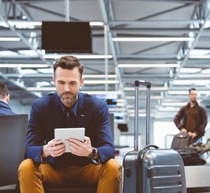 Ein Mann sitzt auf einer Bank im Flughafen und schaut auf sein Tablet. 