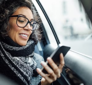 Smiling woman with glasses sitting in a car, using her smartphone to manage invoices on the go.