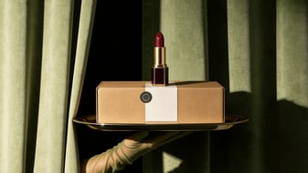 A serving hand holding a tray with a red lipstick on a box in front of green curtains.