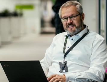 Mitarbeiter mit Brille sitzt mit einem Laptop im Büroflur und blickt in die Kamera