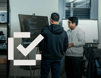 Checklist item marked as complete. Two colleagues collaborating in an office, discussing software development work displayed on a large digital screen. The image captures teamwork, knowledge sharing, and problem-solving in a technical environment.