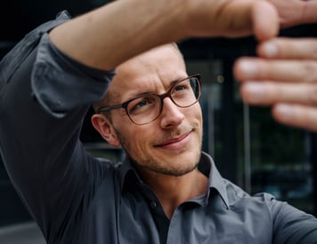 En mann med briller og mørk skjorte ser konsentrert ut og former en ramme med hendene, som om han visualiserer en perspektiv.