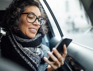 Smilende kvinne med briller sitter i en bil og bruker smarttelefonen sin til å håndtere fakturaer mens hun er på farten.