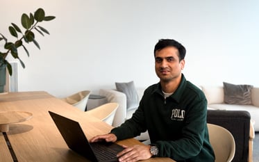 Pankaj, a person with short black hair and light facial hair sits at a long wooden table in a modern office or co-working space. They are wearing a dark green sweater with a white Polo logo and a silver wristwatch. Their hands are resting on a black laptop, and they are smiling gently at the camera. In the background, there are beige chairs, a sofa with cushions, and a large green plant against a white wall.