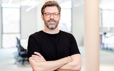 Mathias, a person with short brown-gray hair, a full beard, and brown glasses stands with crossed arms in a modern office. They are wearing a black T-shirt and looking confidently at the camera. In the background, large windows, bright walls, and some office chairs are visible.