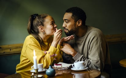 Ein junges Paar sitzt entspannt an einem kleinen Essenstisch und die Frau führt ein Stück Kuchen zu dem Mund von dem Mann