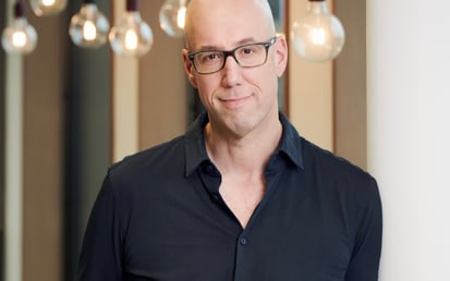 Professional portrait of a man in casual business attire standing in a modern office environment with soft lighting and a blurred background.