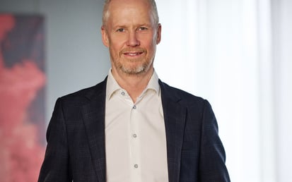 Porträtt av en man i mörk kavaj och vit skjorta, stående inomhus.