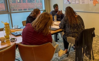 Workshopsessie bij Riverty waarbij deelnemers rond een tafel zitten en luisteren naar een ervaringsdeskundige van Stichting ONSbank, met notities en posters over jongeren en financiën zichtbaar aan de muur in een vergaderruimte.