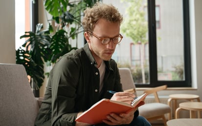 A man with glasses sits on an armchair and writes in a red book