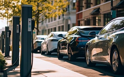 A row of electric vehicles parked at charging stations along a city street on a sunny day, with buildings and trees in the background.