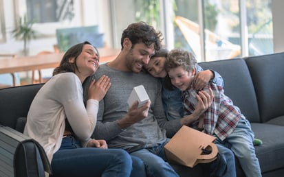 Eine Mama und ein Papa sitzen gemeinsam mit ihrem Sohn und ihrer Tochter auf dem Sofa übergeben Geschenke und umarmen sich