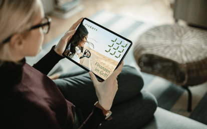 Eine blonde Frau sitzt auf dem Sofa und schaut sich auf ihrem Tablet den Riverty Finanzplaner.