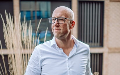 Kale man met een bril en een wit overhemd die buiten staat en bedachtzaam opzij kijkt, met een modern gebouw op de achtergrond