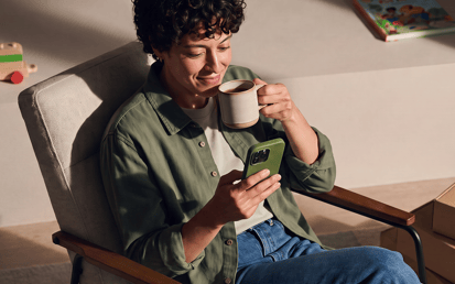 Person sitting relaxed in an armchair, looking at a smartphone and holding a mug.