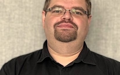 A person is standing in front of a neutral, textured grey background. They are wearing a short-sleeved black button-up shirt and a dark smartwatch on their left wrist. Their arms are loosely folded in front of their body. The person has short brown hair, wears thin-framed metal glasses, and has a neatly trimmed goatee. Their facial expression appears calm and friendly. No branding or accessibility-relevant features are visible in the image.