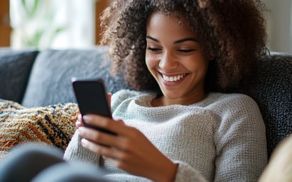 A woman is sitting on the couch, looking at her phone and smiling as she is on her phone.