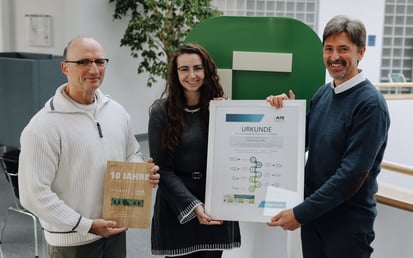 Drei Personen stehen in einem Innenraum und halten zwei Auszeichnungen. Die Person links hält eine Holzplakette zum zehnjährigen Jubiläum der Partnerschaft, während die beiden Personen in der Mitte und rechts eine gerahmte Urkunde präsentieren. Im Hintergrund ist ein großes grünes Riverty-Logo zu sehen.