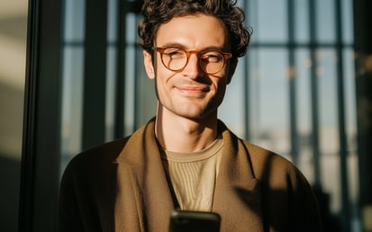A young man using his smartphone in a bright modern office, smiling confidently in the sunlight.