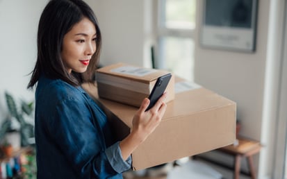 A woman carrying two cardboard packages in her left arm is looking at her smartphone while standing indoors. She is wearing a blue jacket and has a slight smile on her face. In the background, a softly lit room is visible with a blurred desk and a window.