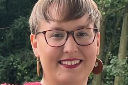 A person with short light brown hair and glasses is smiling outdoors, wearing a textured red top and round earrings, with green trees in the background.