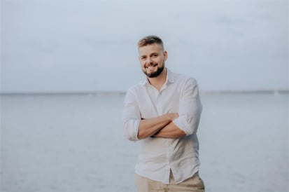 Man with short hair and beard wearing a light linen shirt and beige pants, standing with arms crossed and smiling by the water.