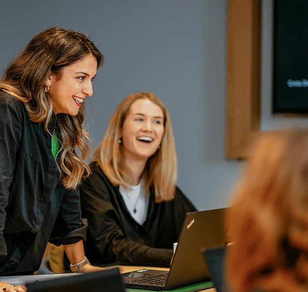 Two smiling colleagues in a meeting room – one leaning over the table, the other seated at her laptop – talking with a colleague in the foreground. A presentation screen hangs on the wall in the background.