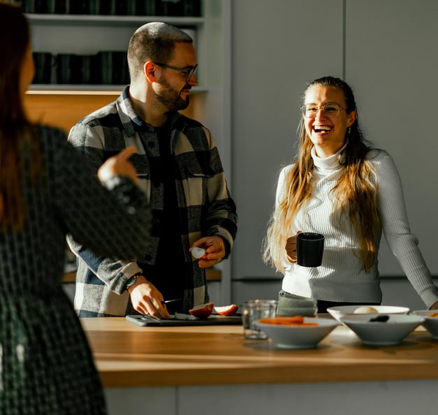 Three people are chatting and laughing in a modern kitchen. One person with long brown hair is standing with their back to the camera, gesturing. Another person with glasses, a beard, and a checkered shirt is cutting fruit and looking towards the first person. The third person, with long hair, glasses, and a white turtleneck sweater, leans against the kitchen island, holding a black mug and laughing. On the countertop, there are bowls of fruit and vegetables, as well as pots with fresh herbs. Sunlight streams through the window, illuminating the scene.