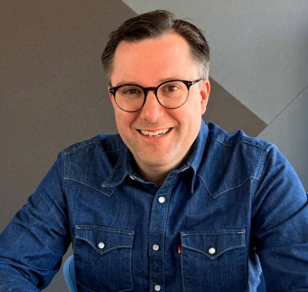 Das Foto zeigt eine Person, die leicht nach vorne geneigt direkt in die Kamera schaut und dabei herzlich lächelt. Die Perspektive wirkt leicht von oben aufgenommen, was dem Bild eine nahbare, persönliche Wirkung verleiht. Die Person trägt eine runde, schwarze Hornbrille mit einem erkennbaren „T"-Logo auf dem Nasensteg, was auf die Brillenmarke Tom Ford hindeutet. Sie trägt ein mittelblau gefärbtes Jeanshemd der Marke Levi's, erkennbar am roten Markenzeichen auf der rechten Brusttasche. Das Hemd hat zwei aufgesetzte Brusttaschen mit weißen Knöpfen und sichtbaren gelben Nähten. Die Person hat kurzes, dunkelbraunes Haar mit leichten grauen Akzenten an den Seiten und Schläfen. Der Gesichtsausdruck wirkt offen, positiv und einladend. Im unteren Bildbereich ist ansatzweise ein blauer Stuhl oder Sitzgegenstand sichtbar. Der Hintergrund besteht aus einer modernen, geometrisch gestalteten Wand in verschiedenen Grautönen. Die Wand zeigt großflächige, diagonale Flächen in Hell und Dunkelgrau, die einen zeitgemäßen, professionellen Raumeindruck erzeugen. Links im Bild ist ein schmaler, heller Streifen zu sehen, der auf eine Wand oder ein Bauelement in Beige oder Hellgrau hindeutet.