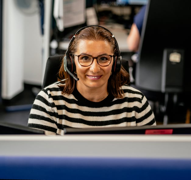A woman with brown hair, wearing glasses and a black-and-white striped sweater, is smiling at the camera while seated at her desk in an office environment. She is wearing a headset. In the background, another person is working at a computer.