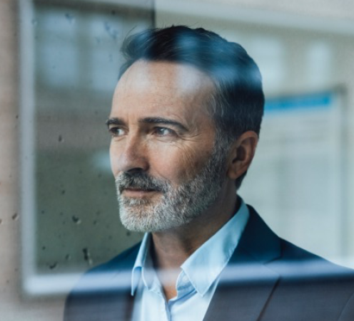 Man in pak met grijze baard kijkt uit het raam, met een gedeeltelijke reflectie in het glas.