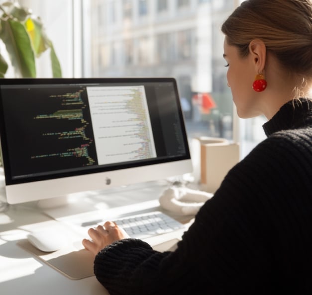 Woman working at an iMac displaying code in a bright office setting, with large window in the background