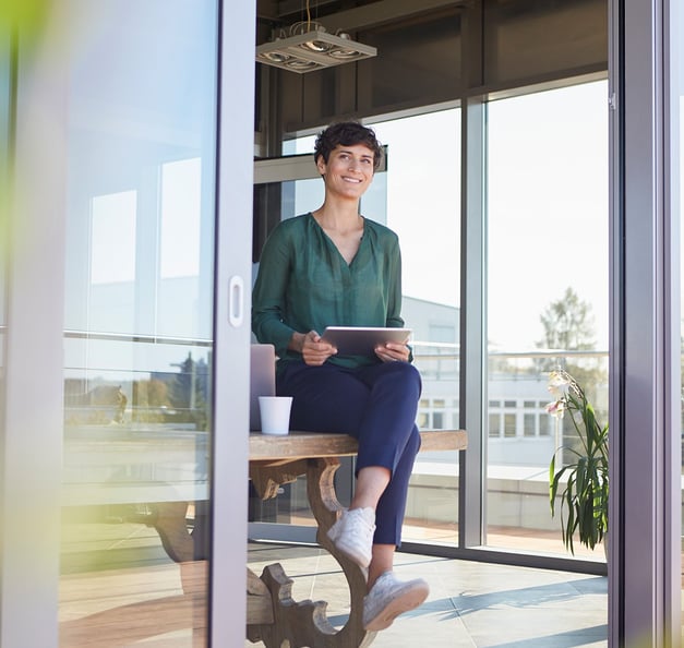 eine Frau in grüner Bluse sitzt an einem Tisch mit einem Tablet in der Hand und einem Computer und einer Tasse Kaffee neben sich.