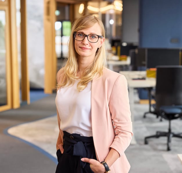 A blonde woman wearing dark-framed glasses, a white top, a light pink blazer, and dark high-waisted tied trousers stands smiling with her hands in her pockets in a spacious, modern office background.
