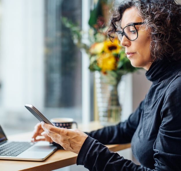 Kvinna, sittande vid laptop med smartphone i handen.