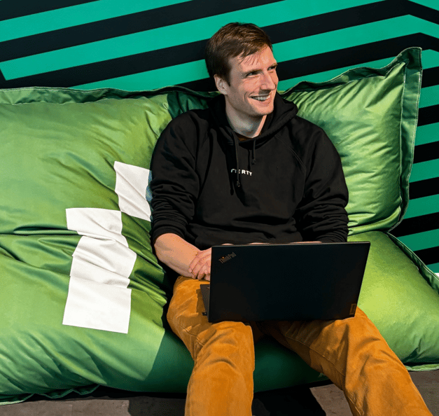 A person is sitting relaxed on a large green beanbag with a white geometric line pattern. The person is wearing a black hoodie and mustard-colored pants. On their lap, the person holds a black Lenovo ThinkPad laptop, with the ThinkPad logo visible on the back of the screen. The person is smiling and looking slightly to the left, outside the frame. The background is made up of diagonal green and black stripes, creating a graphic, modern look. No other people, furniture, or readable text are visible in the image.